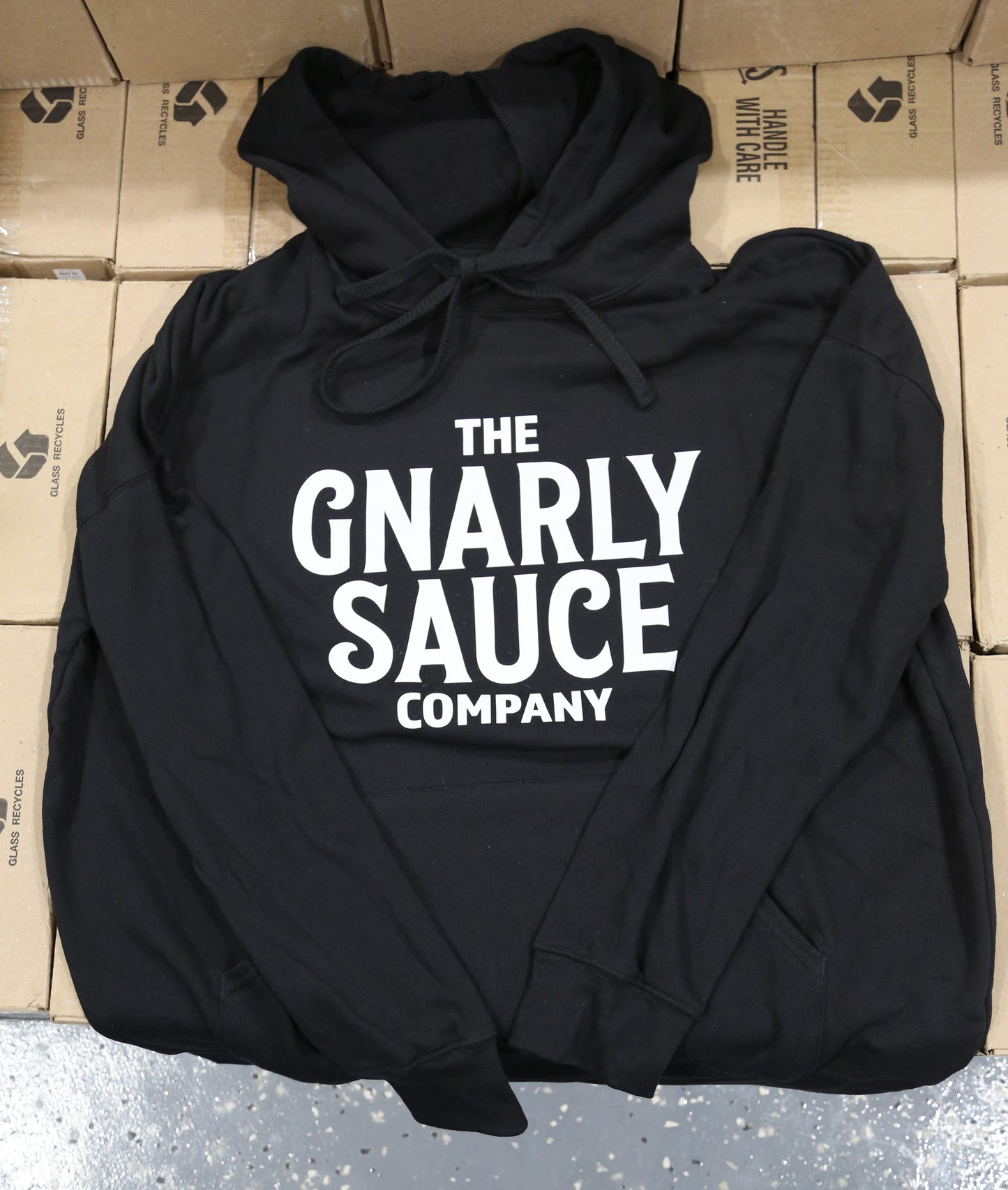 Black hoodie with 'The Gnarly Sauce Company' logo in white on cardboard boxes