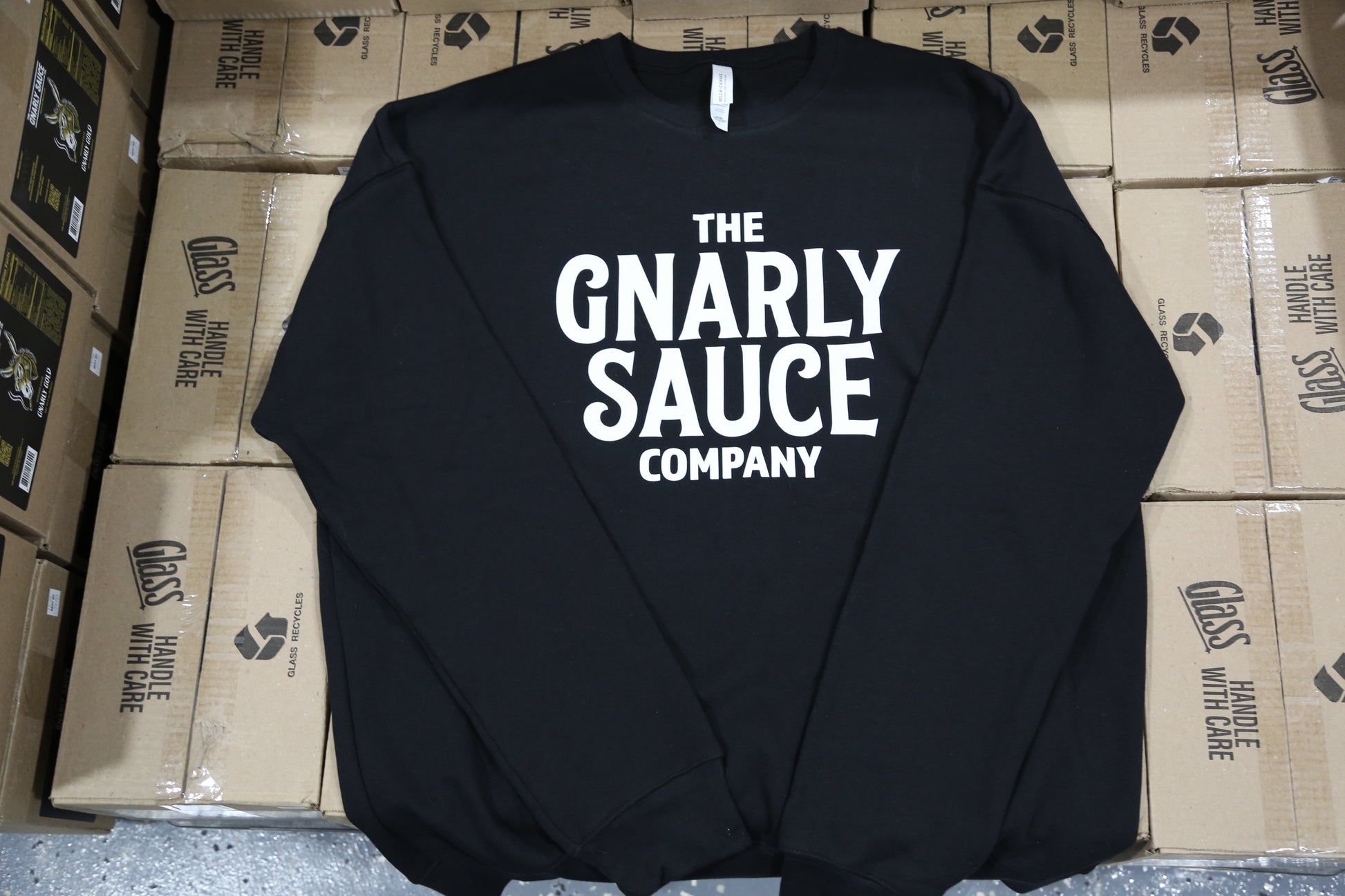 Black sweatshirt with 'The Gnarly Sauce Company' text in white on a background of cardboard boxes.