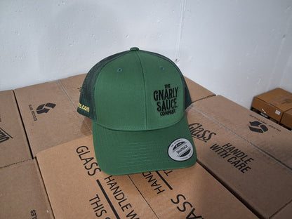Green cap with 'The Gnarly Sauce Company' logo in black sitting on cardboard boxes